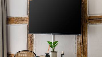 Een kamer met een grote, zwart tv-toestel aan een muur bevestigd, een houten tafel met een bloempot, een afstandsbediening en een kleine fles.