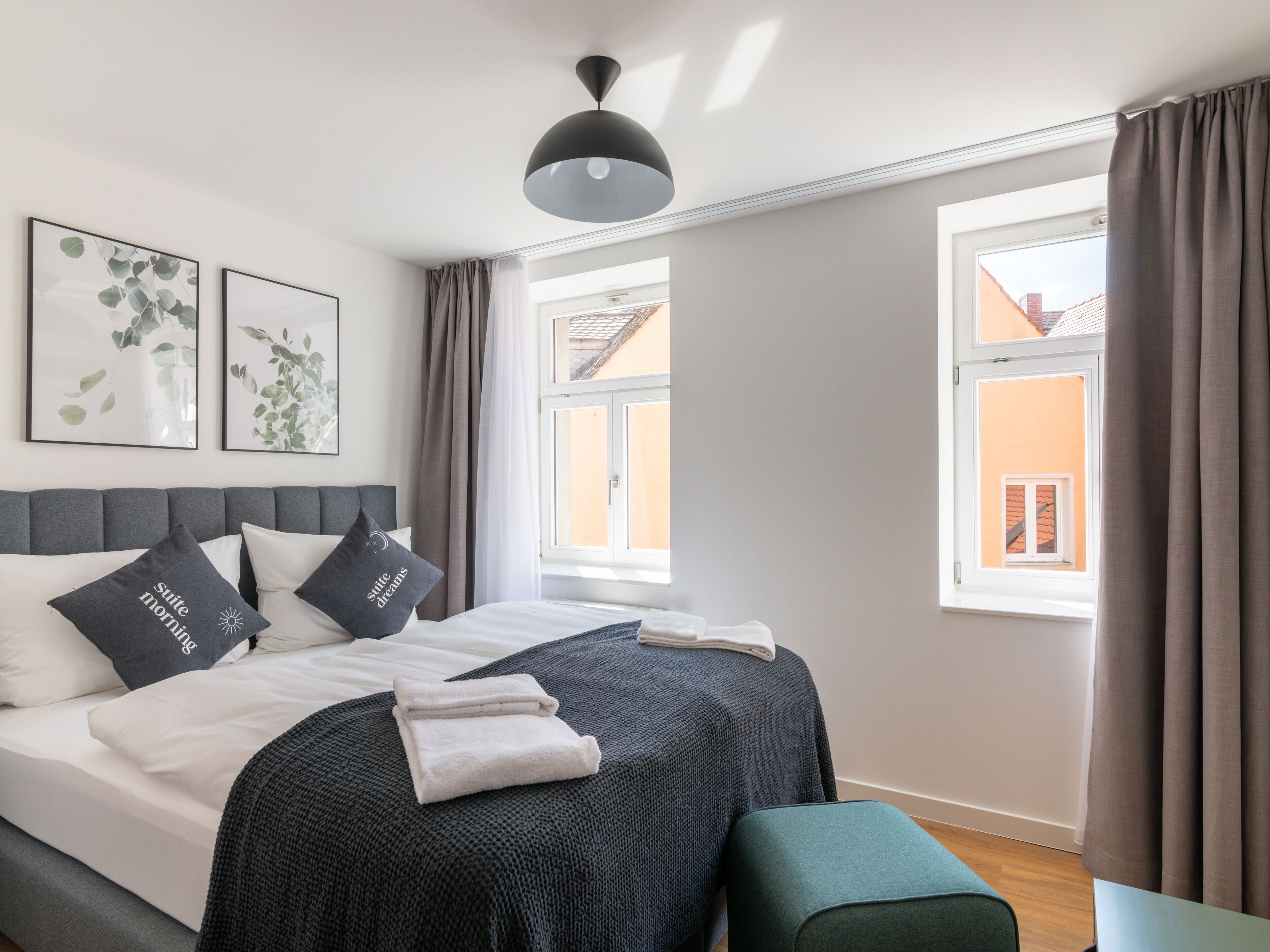 Una cama bien hecha con un cabecero gris, dos almohadas con texto, una manta gris y una toalla doblada. Hay dos ventanas con cortinas, una lámpara en el techo y dos cuadros en la pared.