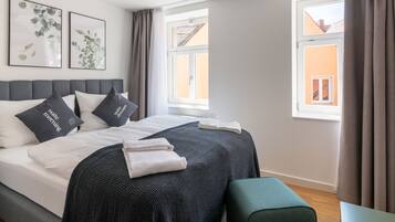 Ein ordentlich bezogenes Bett mit grauem Kopfteil, zwei Kissen mit Aufdruck, eine graue Decke und ein gefaltetes Handtuch. Zwei Fenster mit Vorhängen, eine Deckenleuchte und zwei gerahmte Bilder an der Wand.