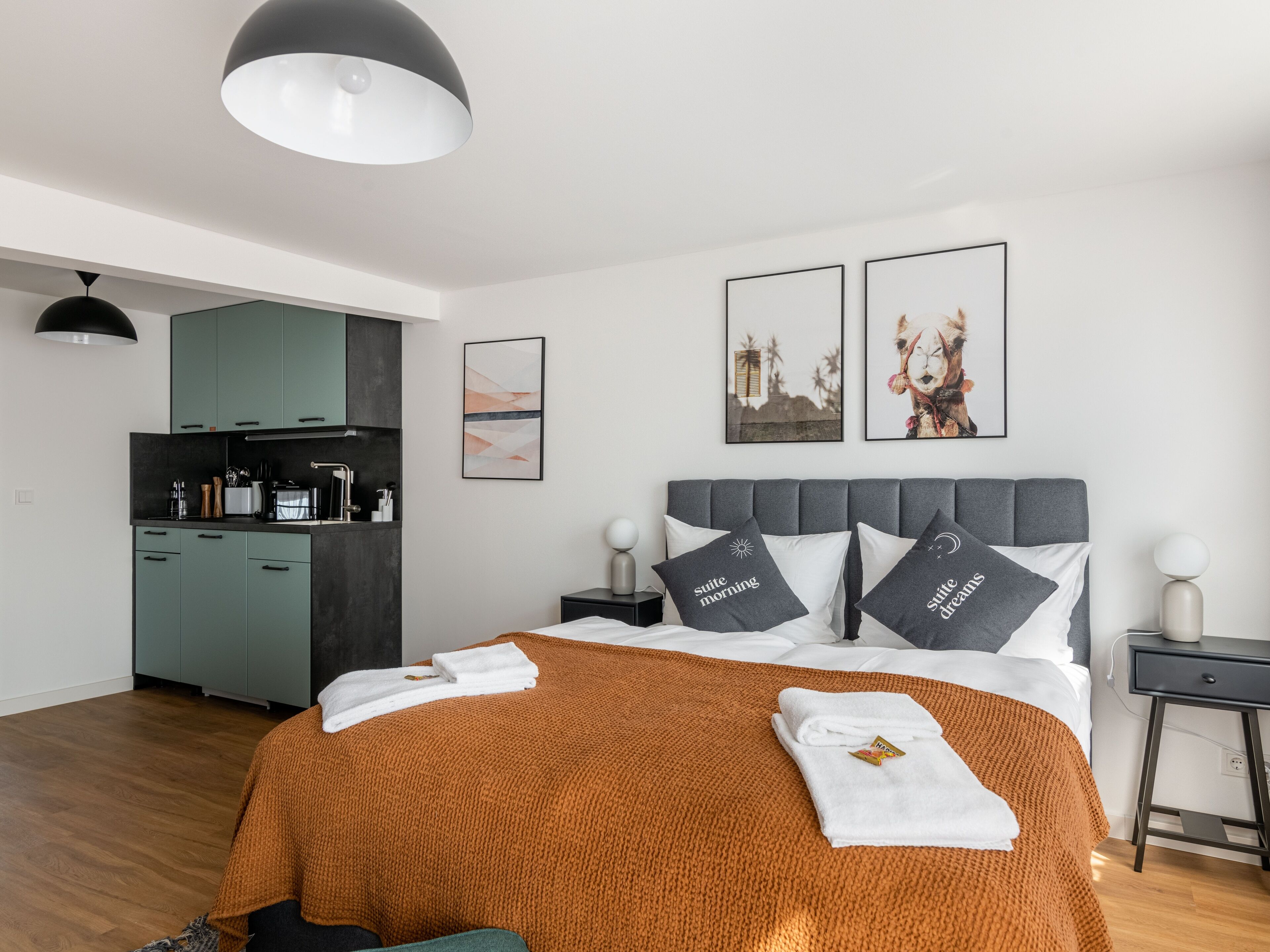 A modern hotel room with a bed, a nightstand, a desk, a kitchenette, and artwork on the walls.