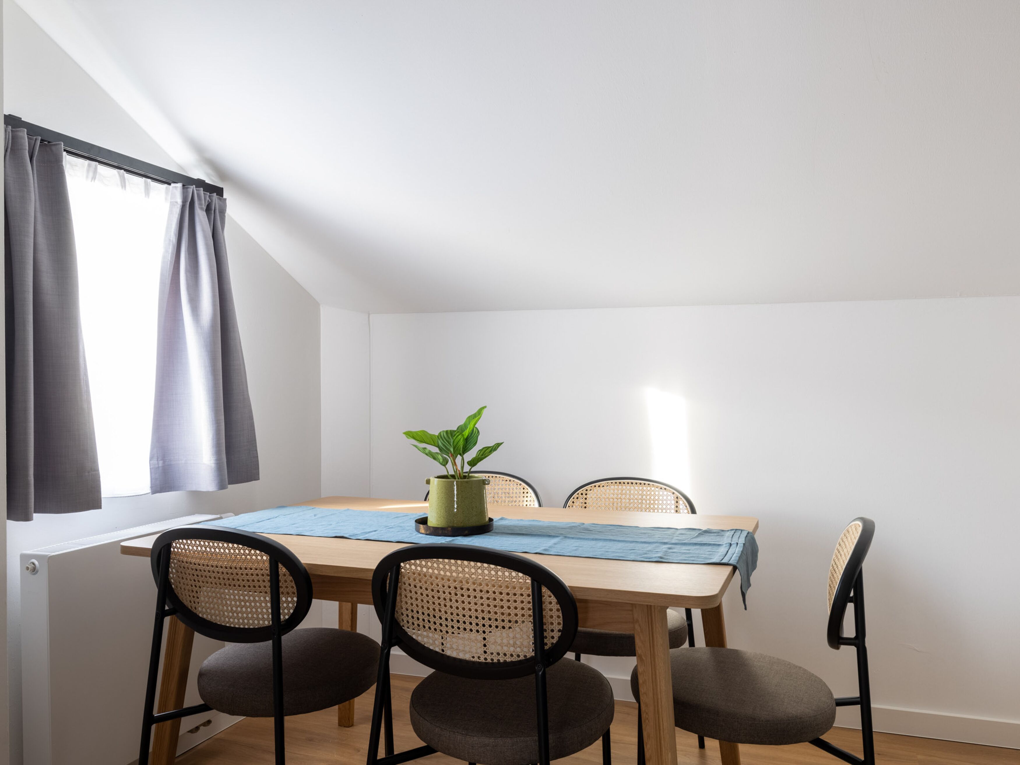 A dining area with a wooden table, four chairs, a potted plant, and a window with curtains.