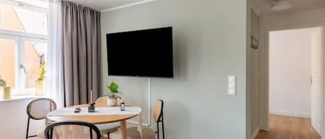 Una sala moderna con un televisor de pantalla plana montado en la pared, una mesa de comedor redonda de madera con cuatro sillas y una planta en el alféizar de la ventana.