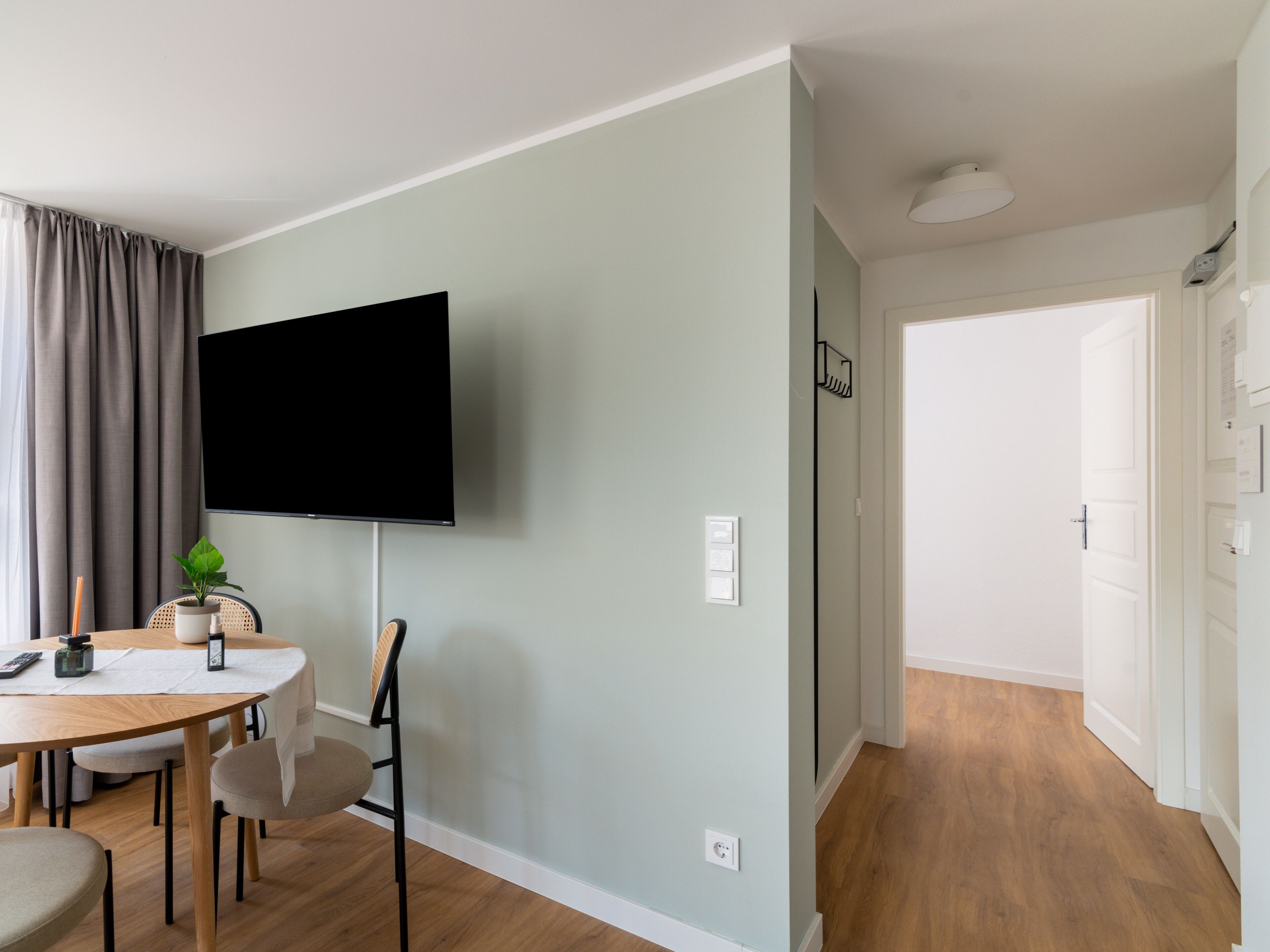 Een moderne woonkamer met een flatscreen tv aan de muur, een eettafel met stoelen, een kleine potplant en een witte deur die naar een andere kamer leidt.