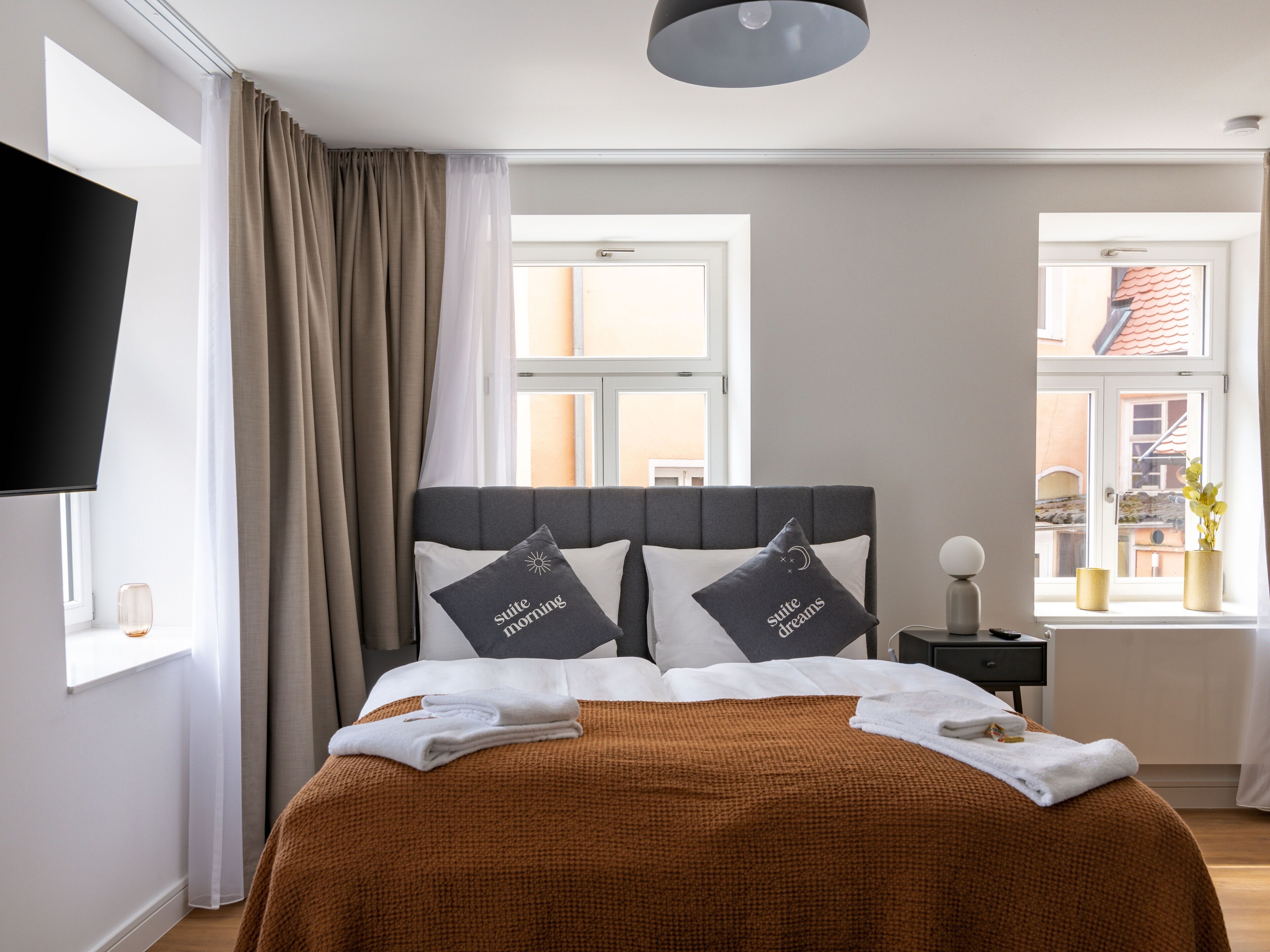 A hotel room with a bed, a TV, a window with curtains, and a bedside table with a lamp.