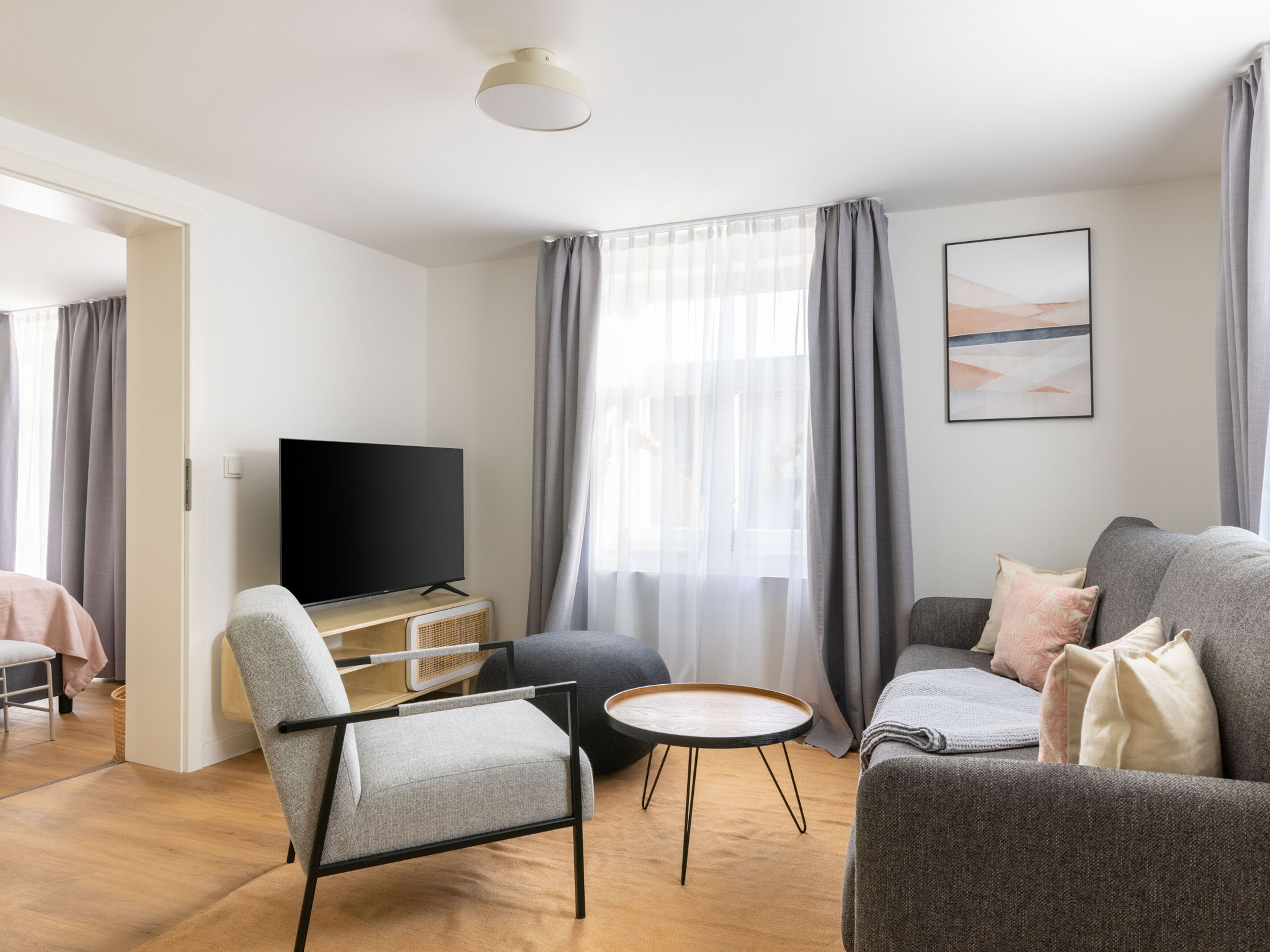 A modern living room with a flat-screen TV, grey sofas, a round coffee table, and a large window with curtains.