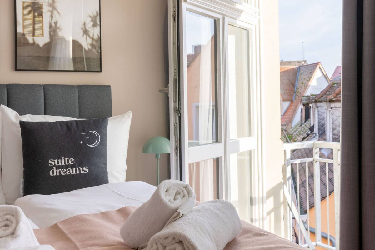 A hotel room with a bed, pillows, a framed picture, and a window with a view of buildings.