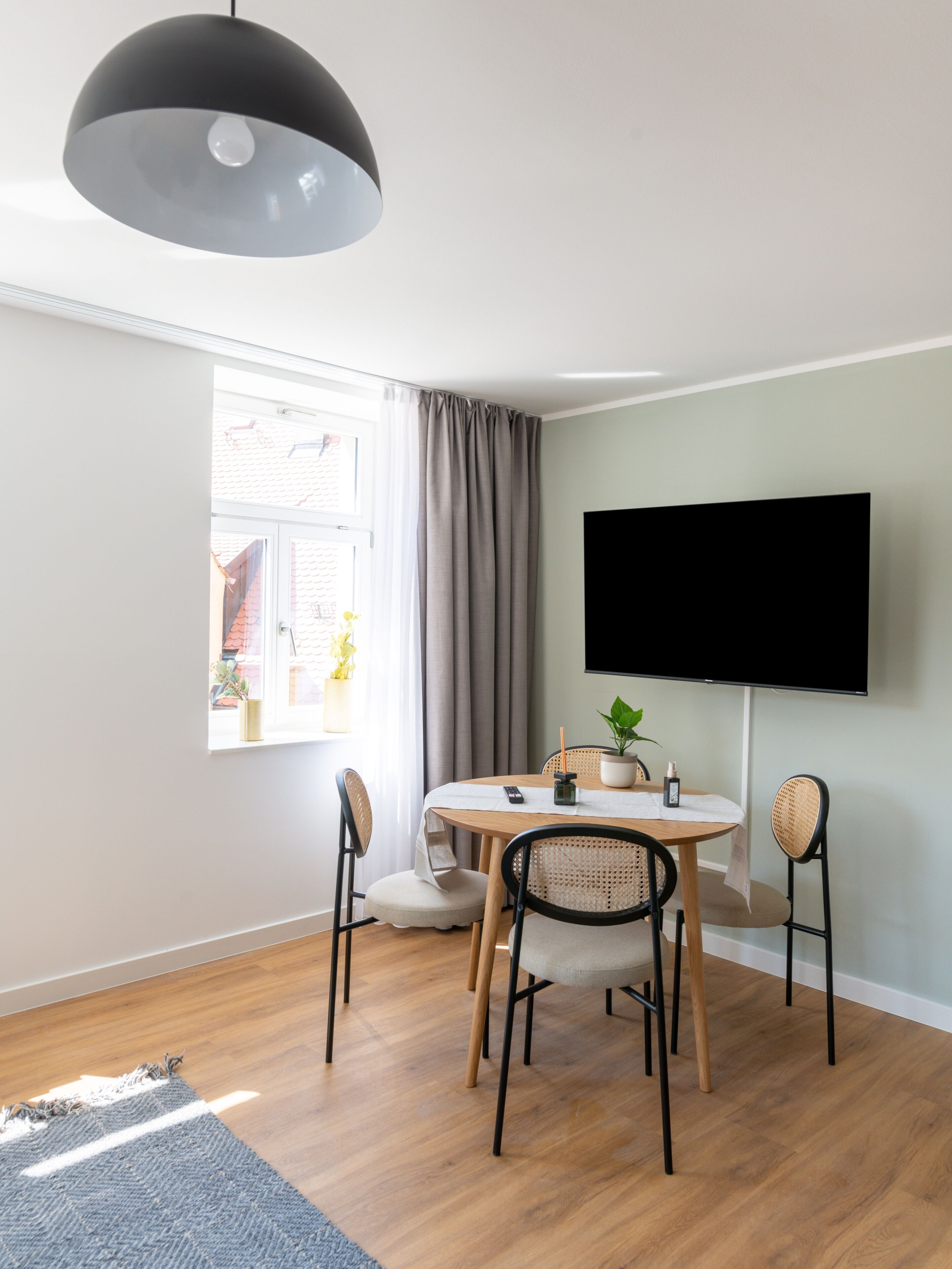 A modern dining area with a round table, four chairs, a potted plant, a remote control, and a flat-screen TV mounted on the wall.