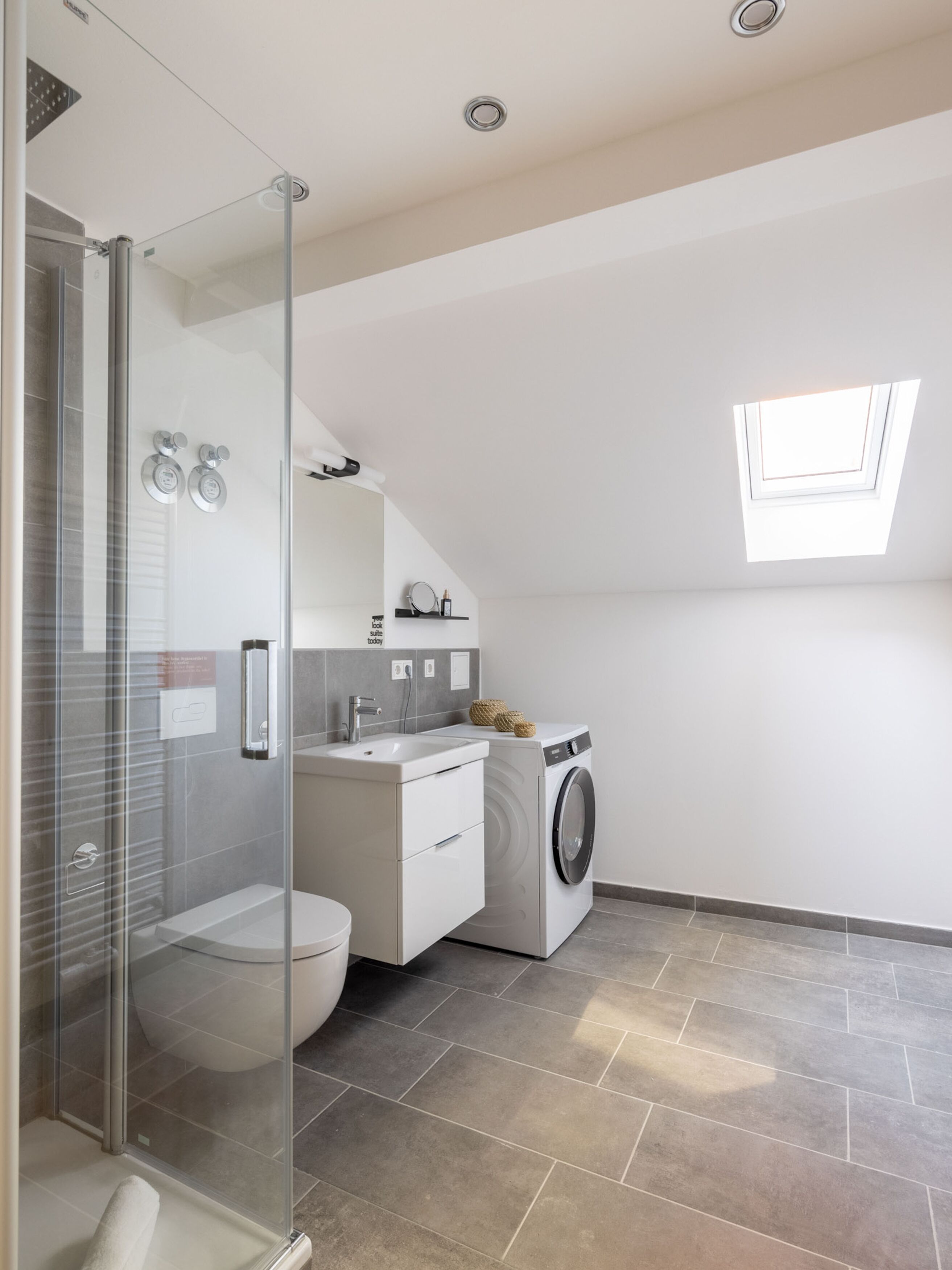 Ein modernes Badezimmer mit einer Duschkabine aus Glas, einer Toilette, einem Waschbecken, einer Waschmaschine und einem kleinen Fenster für natürliches Licht.