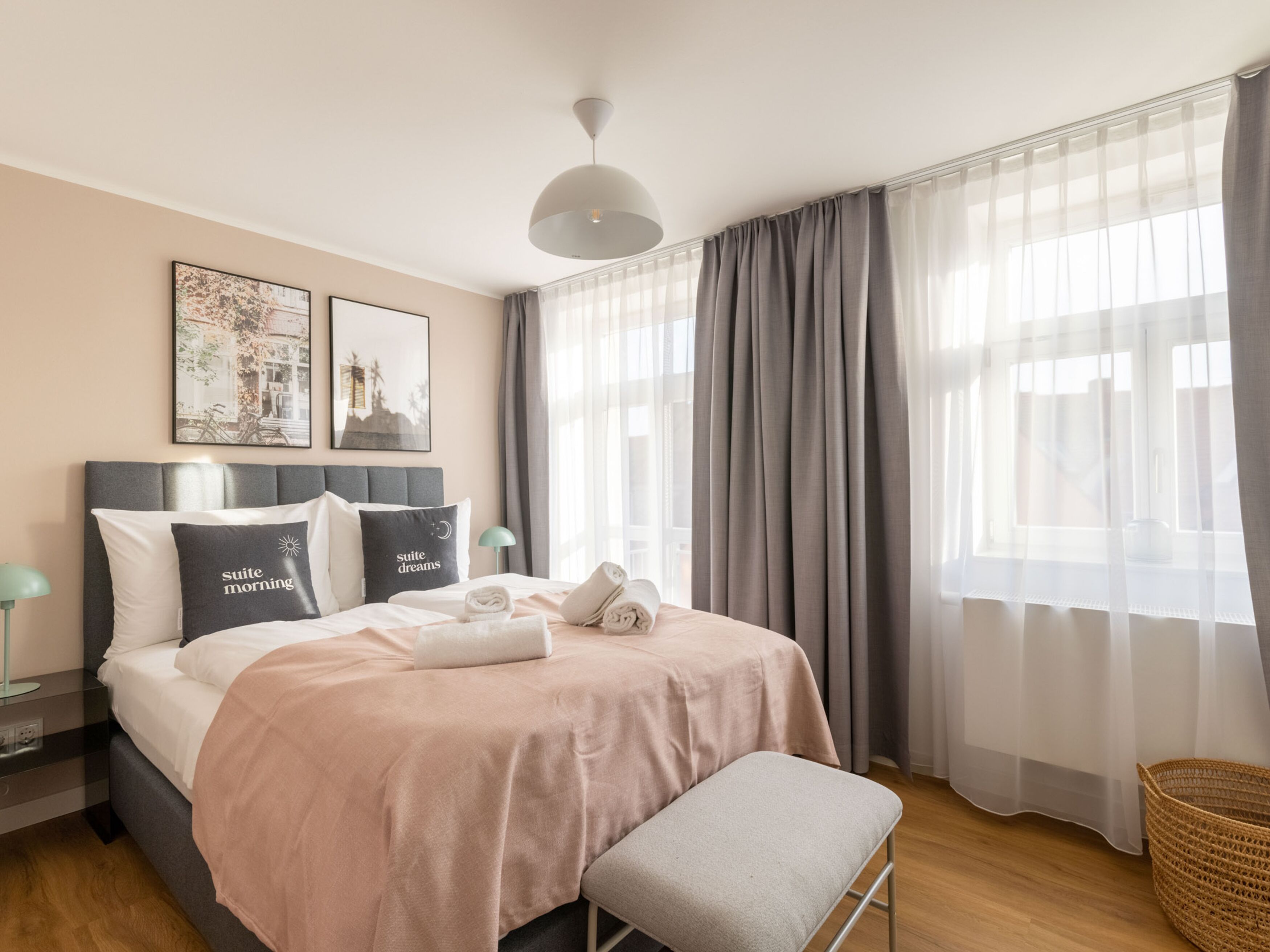 A modern hotel room with a neatly made bed, a bedside table with a lamp, and a large window with sheer curtains.