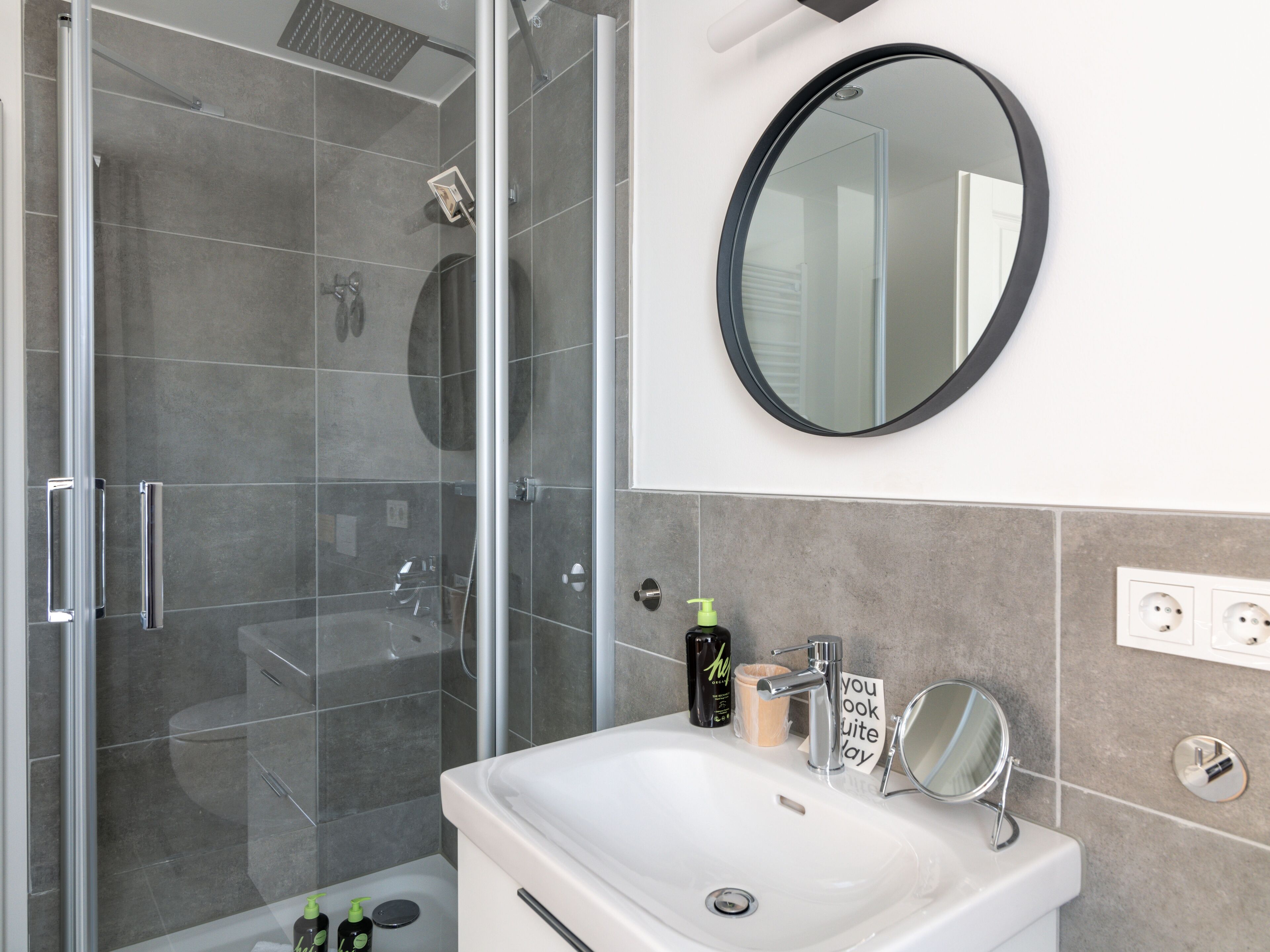 Een moderne badkamer met een douchecabine, een witte wastafel met spiegel en toiletartikelen op het aanrecht.