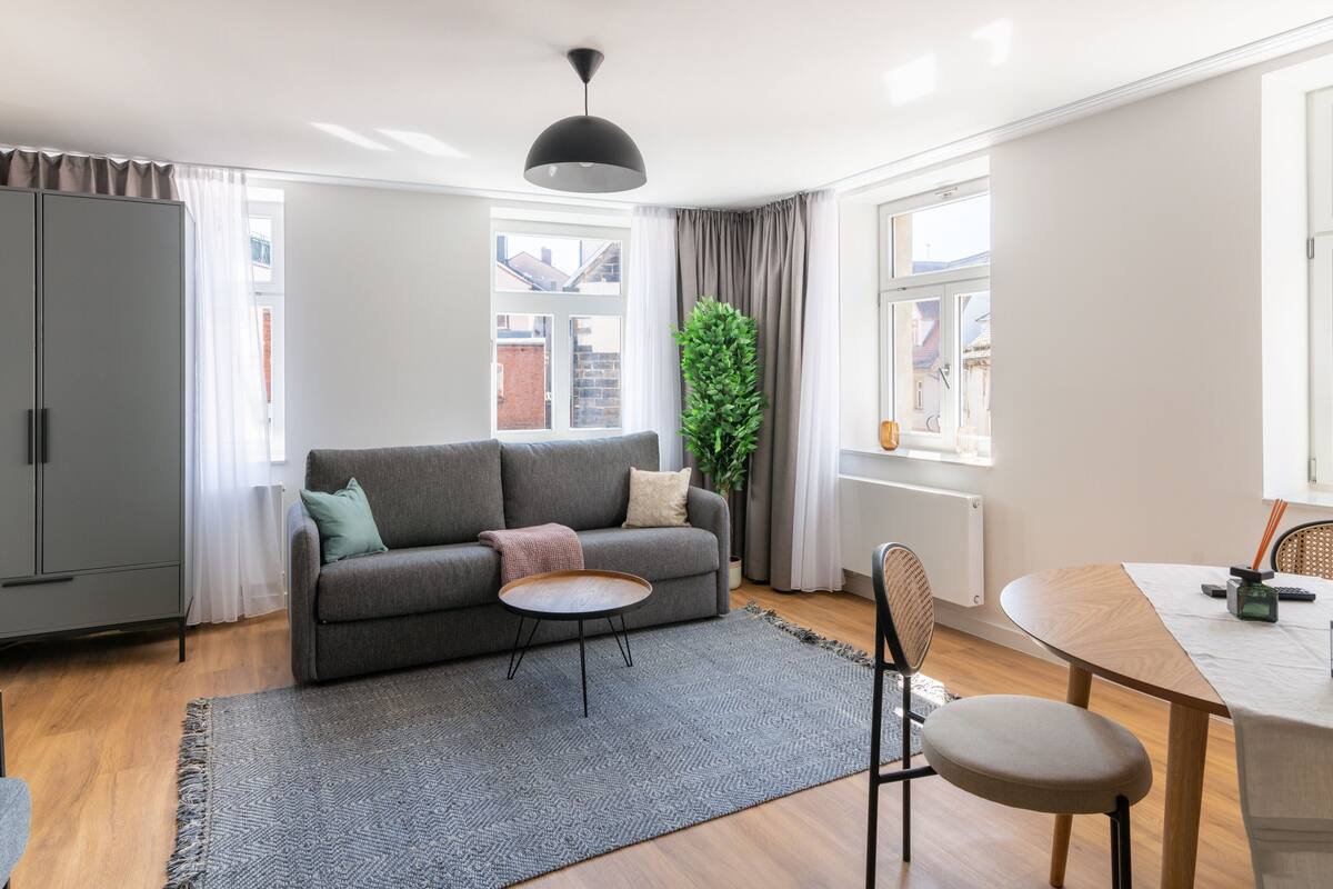 A modern living room with a grey sofa, a small round table, a dining table with chairs, a potted plant, and a large window with curtains.