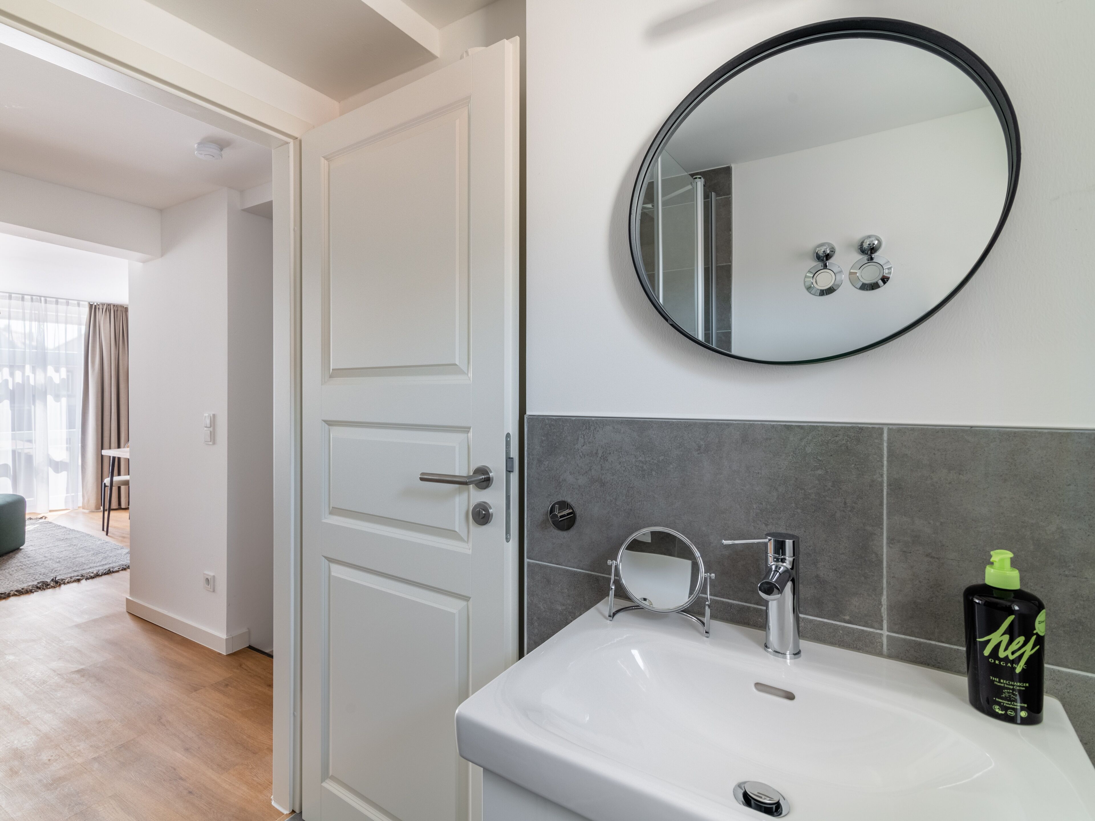 Un baño con un lavamanos blanco, un espejo redondo y una botella de champú orgánico.