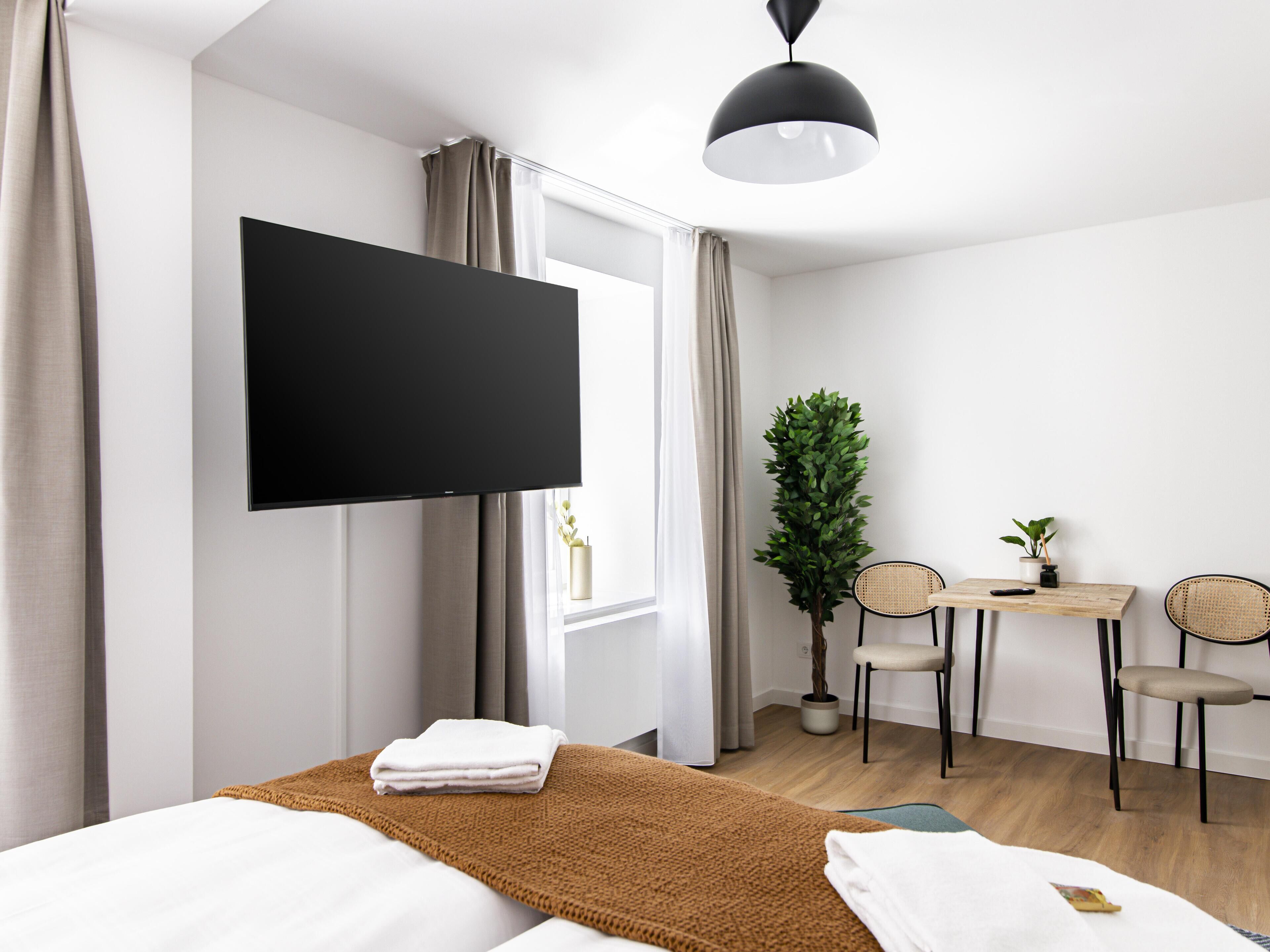 A modern hotel room with a large bed, a flat-screen TV mounted on the wall, a small desk with two chairs, a potted plant, and a window with curtains.