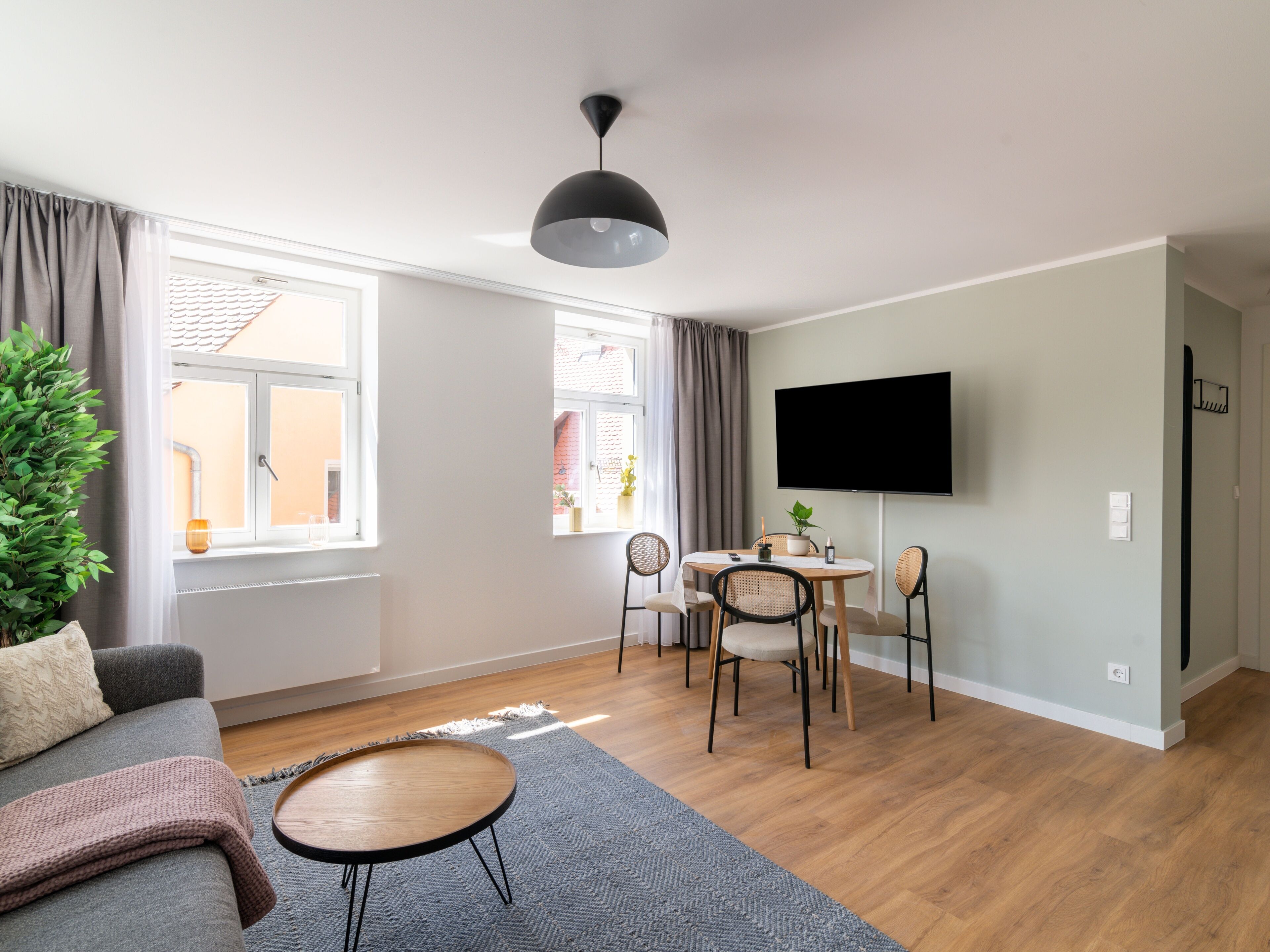 Een moderne woonkamer met een bank, een salontafel, een eettafel met stoelen, een aan de muur bevestigde televisie en een groot raam met gordijnen.
