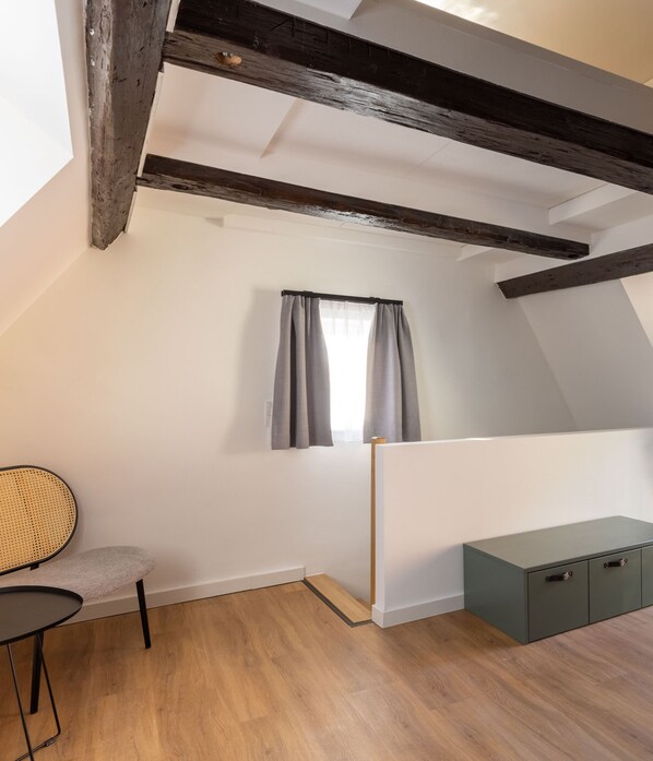 A room with a sloped ceiling, wooden beams, a window with grey curtains, a chair, a small table, and a green cabinet.