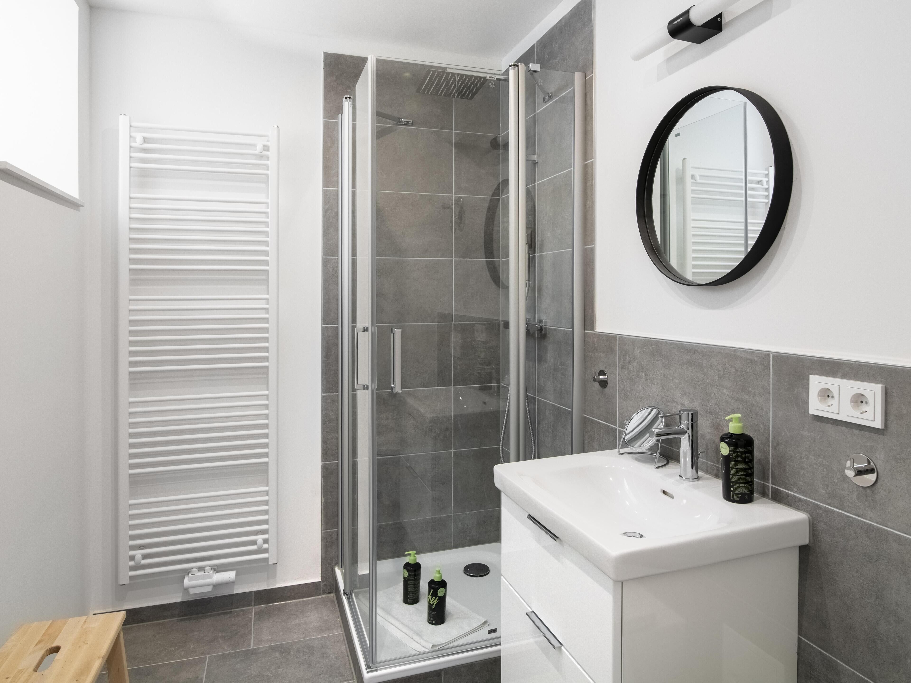 Un baño moderno con ducha de vidrio, un lavabo blanco con espejo y un taburete de madera.