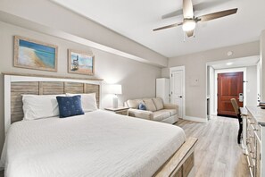 A hotel room with a large bed, a ceiling fan, a desk, and a door leading to another room.