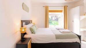 Ein Schlafzimmer mit einem Bett, einer Nachttischlampe, Vorhängen und Blick ins Freie.