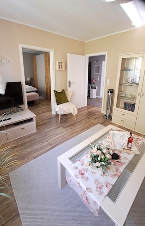 A modern living room with a flat-screen TV, a sofa, a coffee table with a floral tablecloth, and a cabinet with glass doors.
