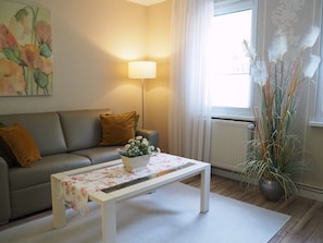 A living room with a sofa, a coffee table with a floral tablecloth, a lamp, and a window with curtains. - Apartment Schlossblick - Top location (Idar-Oberstein)