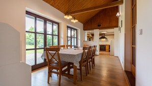 A spacious dining area with a wooden table and chairs, large windows, and a kitchen in the background. - Haus mitten in den Weinbergen - Haus 44. Frühstücksservice möglich! (Liběšice)