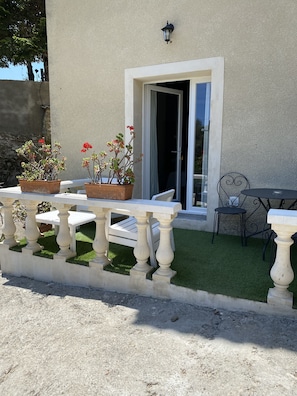 Eine Terrasse mit weißem Tisch und Stühlen, ein kleiner Garten mit künstlichem Rasen und ein Gebäude mit weißer Tür und Fenster.