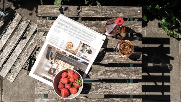 A wooden bench with a magazine, a glass of wine, and a bowl of strawberries.