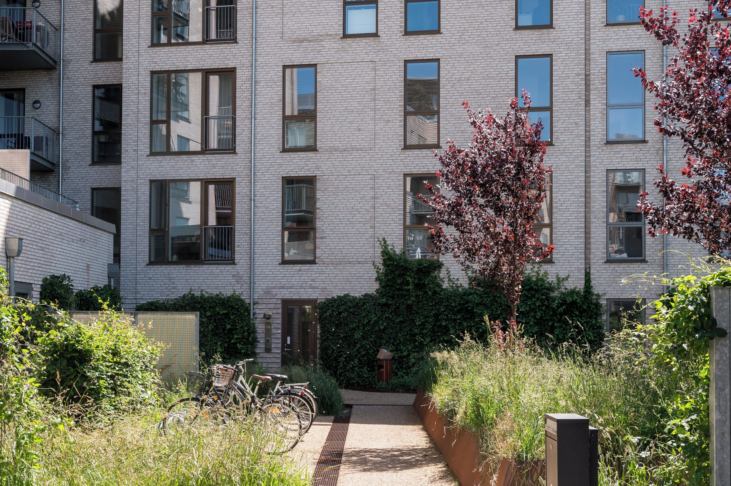 A multi-story residential building with a brick facade, multiple windows, and a small garden area with a pathway and bicycles.