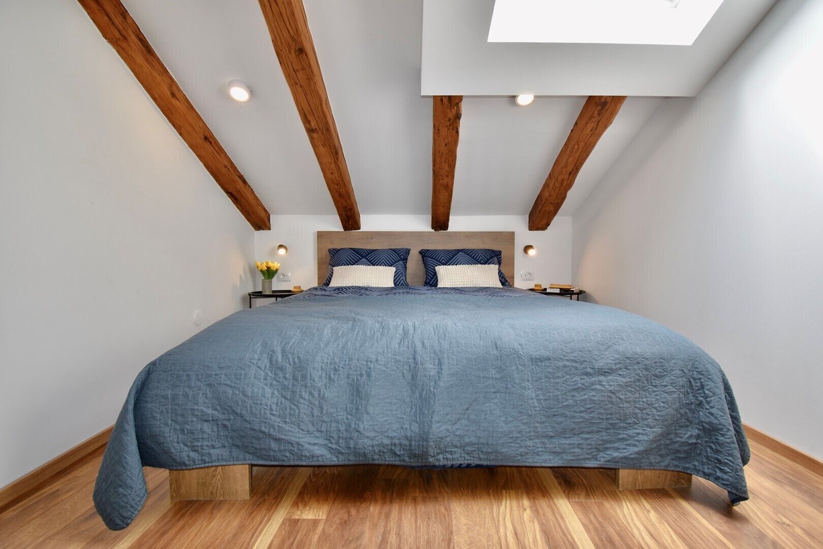 Un dormitorio con una cama grande, vigas de madera, un tragaluz y una mesita de noche con lámpara.
