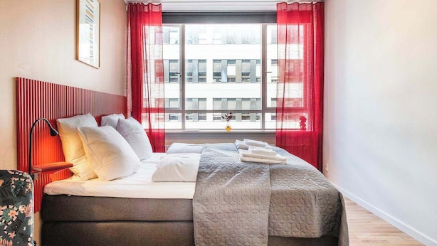 Une chambre d’hôtel moderne avec un grand lit, des rideaux rouges, une vue sur les immeubles et un tableau encadré au mur.