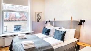 A neatly made bed with white and dark pillows, a grey headboard, and a window with a view of a building with red roofs.