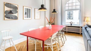 A dining area with a red table and white chairs, framed artwork on the wall, and a window with curtains.