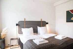 Un letto rifatto con cura, con una testiera grigia, cuscini bianchi e asciugamani piegati su un copriletto marrone scuro.