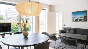 A modern living room with a round dining table, a sofa, and a large window with a view of greenery and buildings. - Beach gardens 2BR by Daniel & Jacob's (Copenhagen)