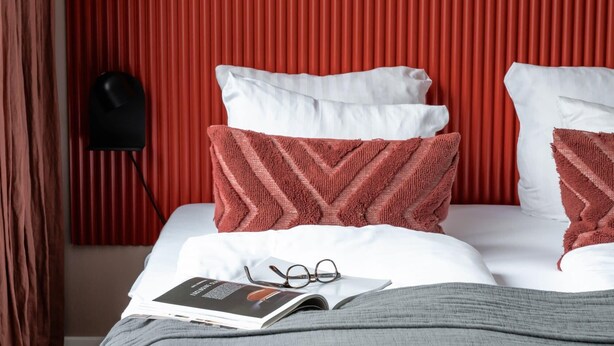 A neatly made bed with white and red pillows, a magazine, and glasses on the bed.