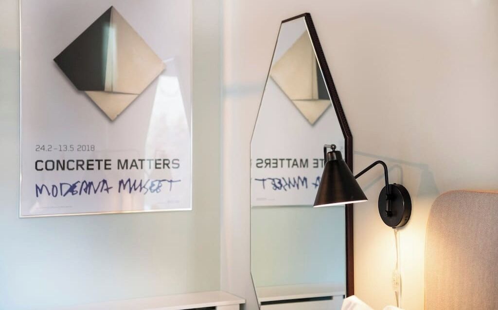 A modern hotel room with a wall-mounted mirror reflecting a geometric artwork titled 'CONCRETE MATTERS MODERN ART MUSEUM' and a black desk lamp.