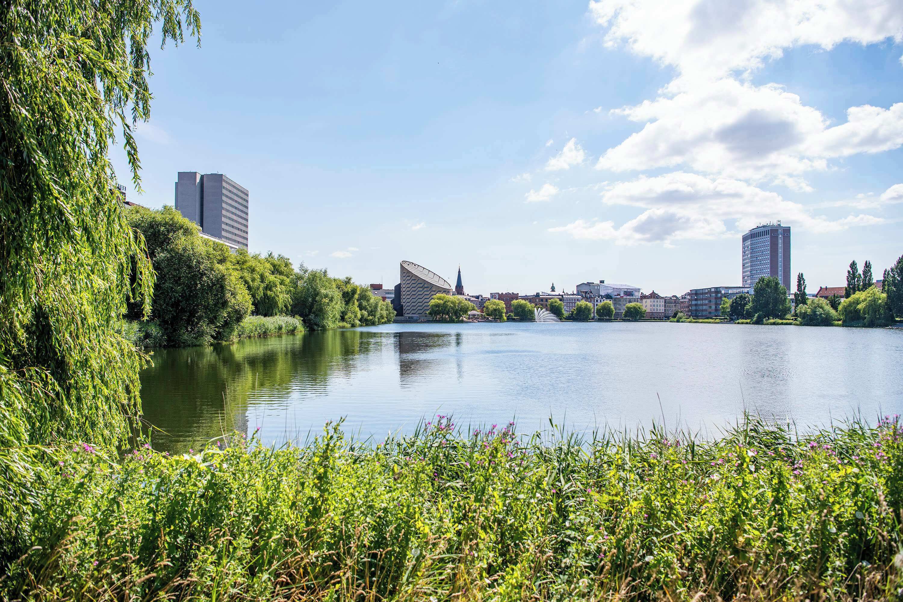 A river with buildings on the opposite bank, surrounded by greenery.