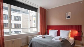 Une chambre d’hôtel avec une grande fenêtre, un lit recouvert de draps blancs, une tête de lit rouge et un radiateur placé sous la fenêtre.