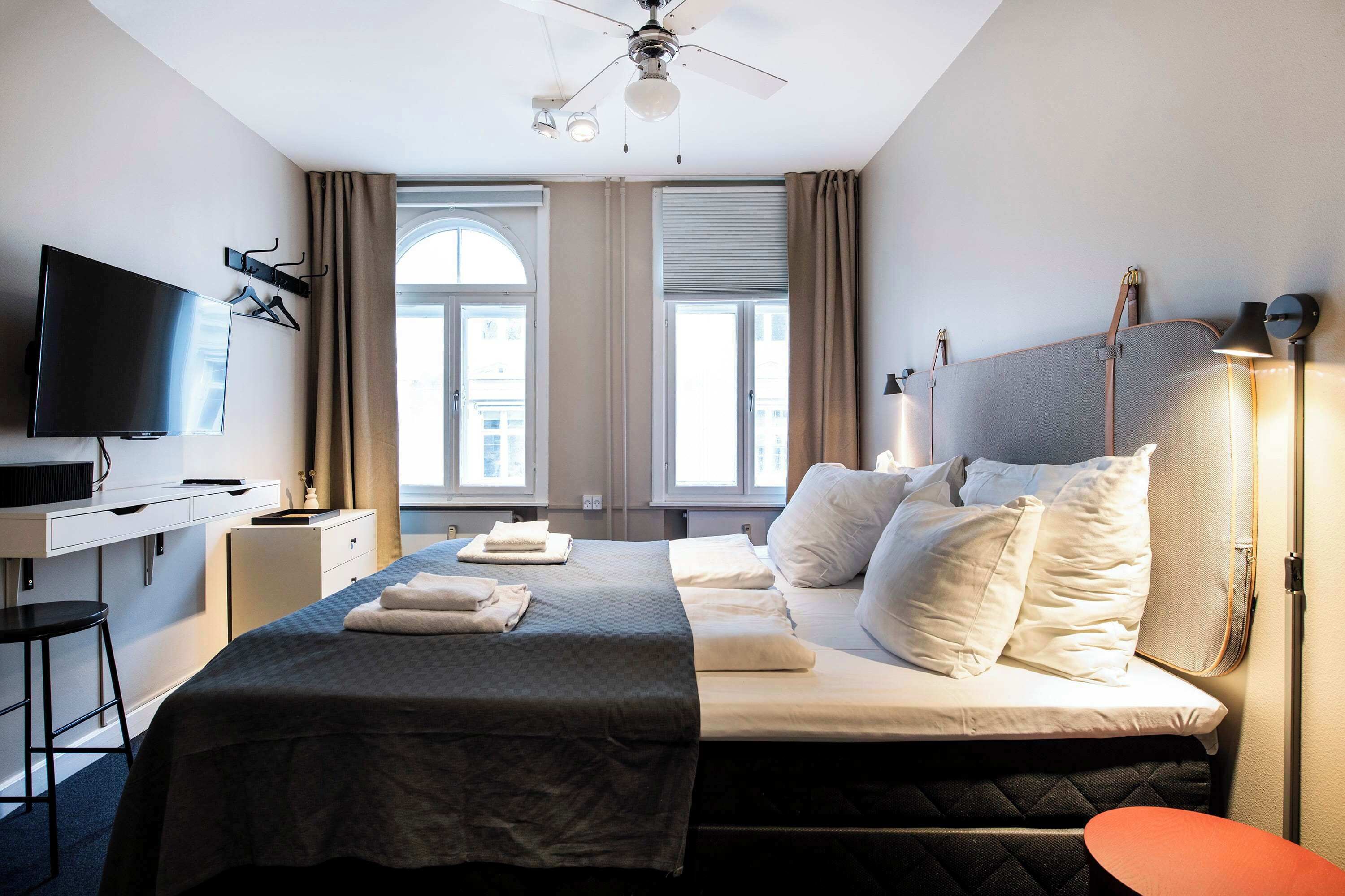 Ett hotellrum med en säng, ett skrivbord med en tv, en takfläkt och stora fönster med gardiner.