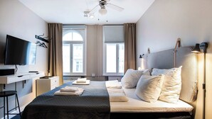 Hotellihuone, jossa on sänky, työpöytä, jossa on televisio, kattotuuletin ja suuret ikkunat, joissa on verhot.