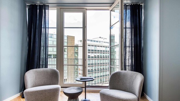 A modern room with two grey armchairs, a small round table, and a view of a cityscape through large windows. - The Lakes 1BR swanky by Daniel & Jacob's (København)