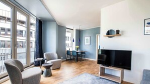 A modern living room with a flat-screen TV, grey armchairs, a dining area with a table and chairs, and large windows with a view of buildings. - The Lakes 1BR swanky by Daniel & Jacob's (København)