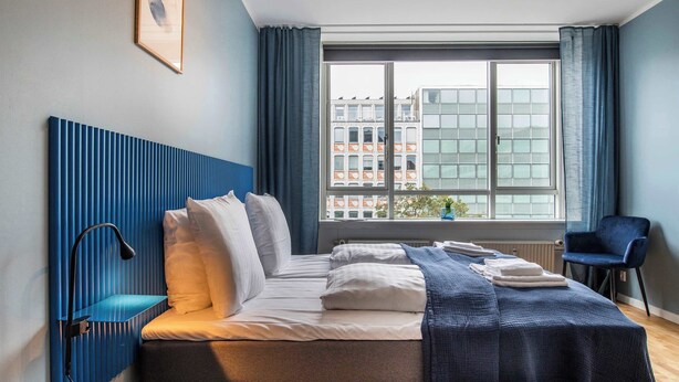A modern hotel room with a large bed, a bedside table, a chair, and a window with a city view.