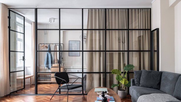 A modern living room with a grey sofa, a wooden coffee table, and a black chair. A large window with curtains is visible, and a plant is placed on the right side. - The Brewer apartments by Daniel&Jacob's (København)