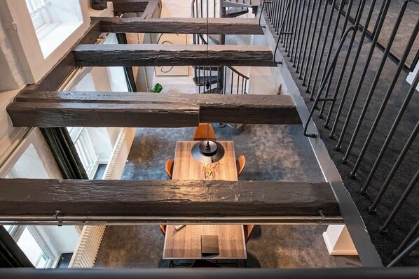 A high-ceilinged interior with exposed wooden beams, a central wooden table with chairs, and a staircase with metal railings.