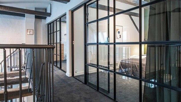 Ein modernes Interieur mit einer Glastrennwand, einer Treppe und einem sichtbaren Bett durch die Trennwand hindurch.
