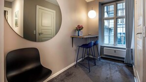 En hall med en rund spegel, en svart stol, ett litet bord med en vas med blommor och ett fönster med gardiner.