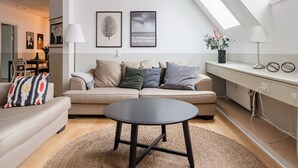 En moderne stue med sofa, rundt sofabord og spiseområde med stole. Tavler med billeder hænger på væggen, der er en lampe, og der står en plante på en hylde.