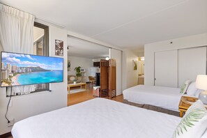 Ein Hotelzimmer mit zwei Betten, einem Fernseher, der eine Stadtansicht zeigt, und Blick auf einen Strand.