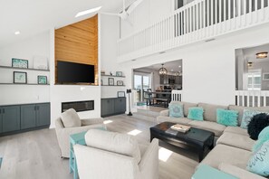 A spacious living room with a high ceiling, a flat-screen TV mounted on a wooden wall, a dining area with a bench, and a kitchen visible through an open door. - 4 bdrm, Beach Front, Sun-Sun Pet Friendly (Cape San Blas)